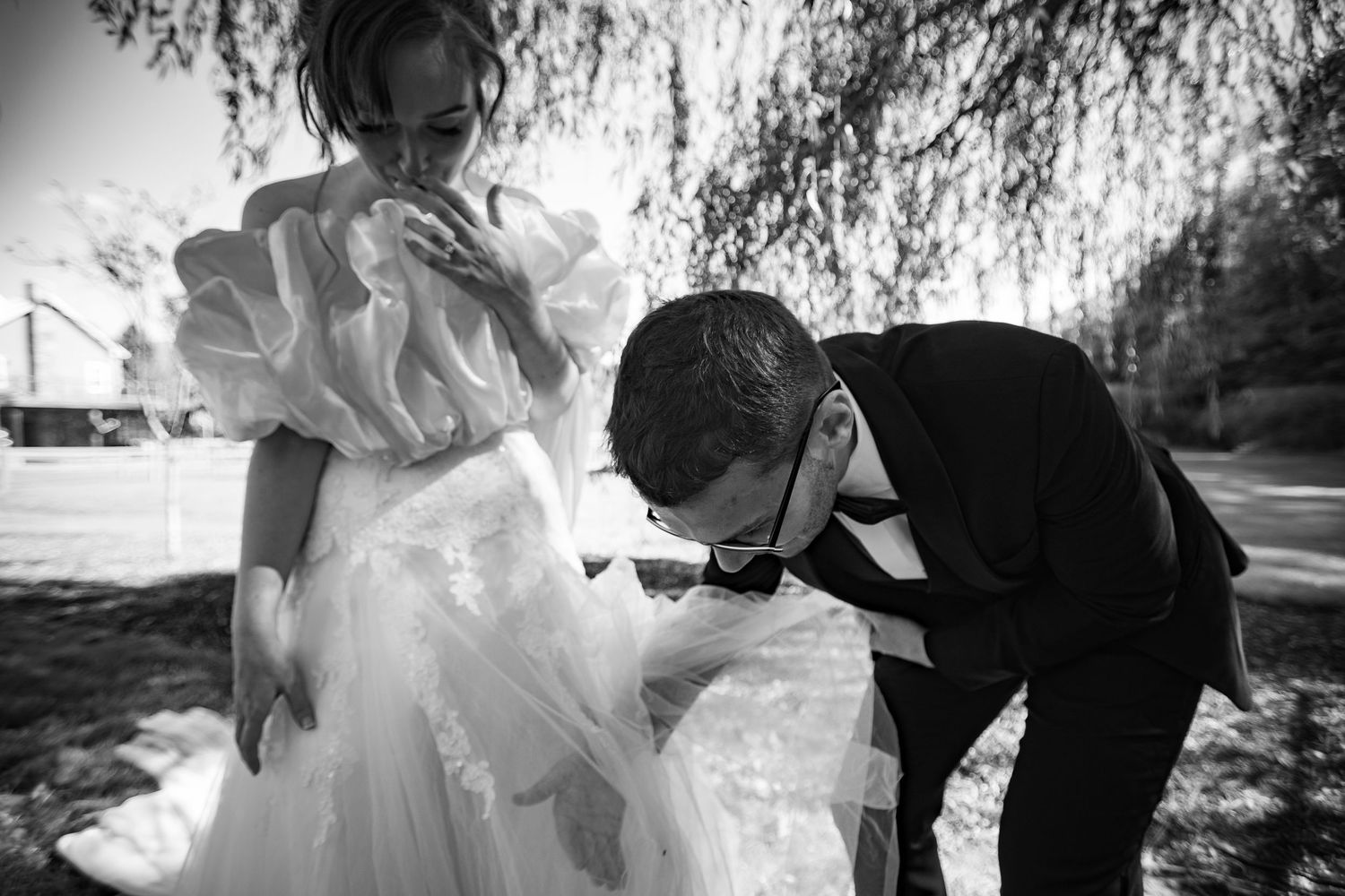 Black and white wedding photo of groom kissing bride's hand outdoors under a willow tree.