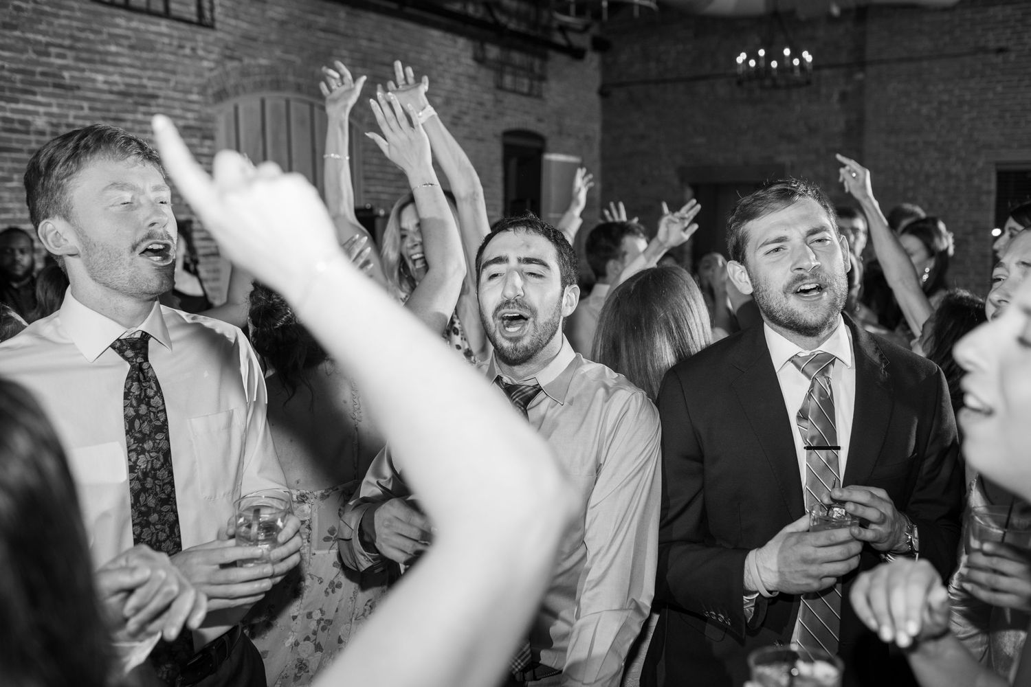 Black and white photo of energetic wedding guests dancing and singing with hands raised at reception.