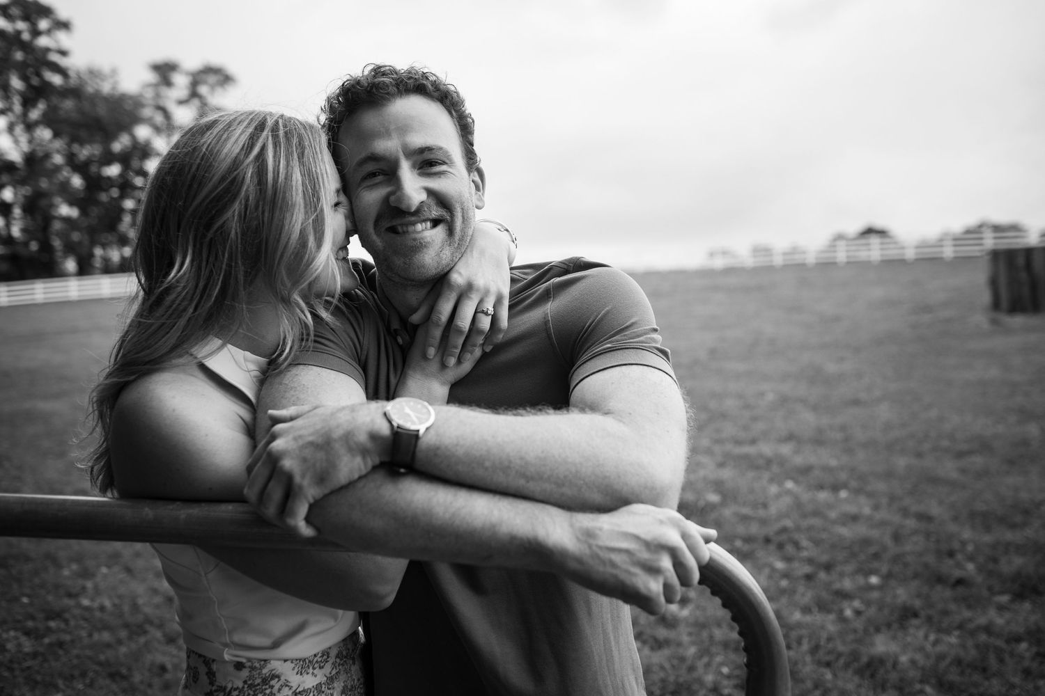 A woman lovingly kisses a smiling man on the cheek outdoors in a black and white romantic couple photo.