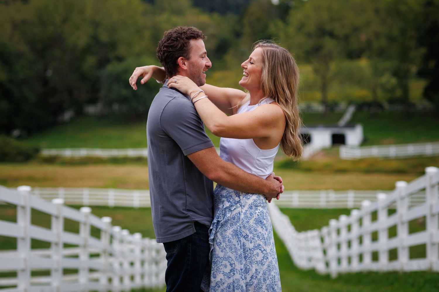 A happy couple embraces and laughs together outdoors on a green farm with white fences and a barn in the background.