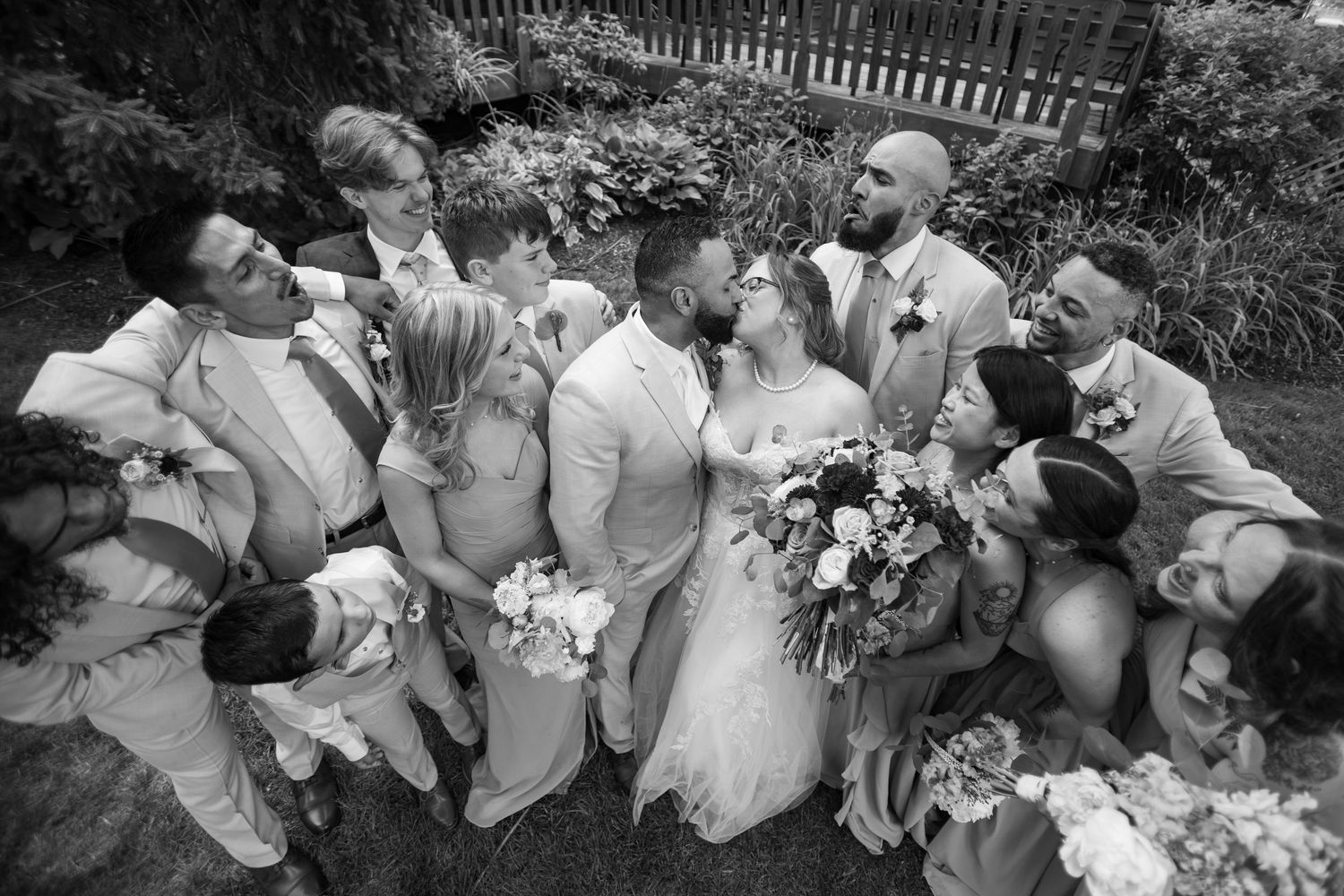 Black and white aerial view of bride, groom, and wedding party gathered closely together in a garden celebration.