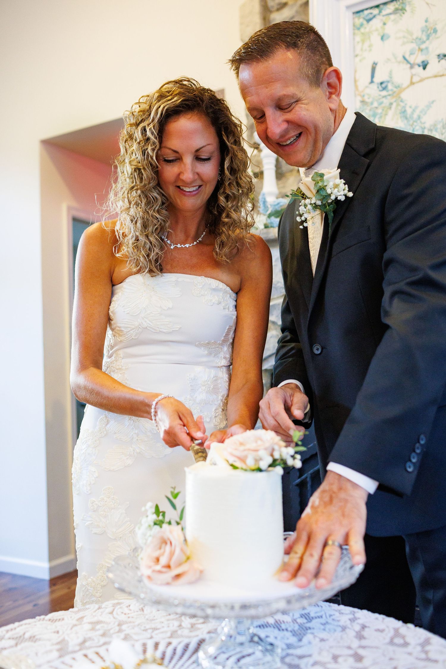 Bride and groom smile while cutting a small white wedding cake decorated with pink flowers.