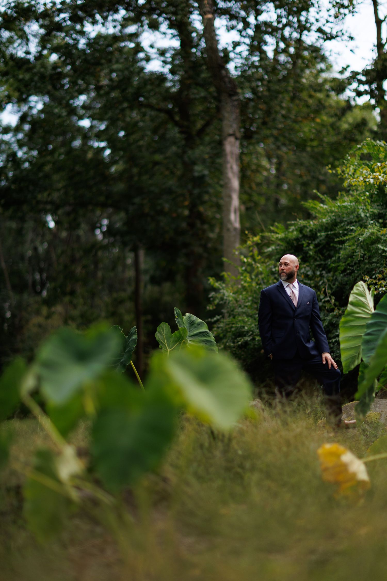 Outdoor wedding scene through foliage with suited figures standing among trees.