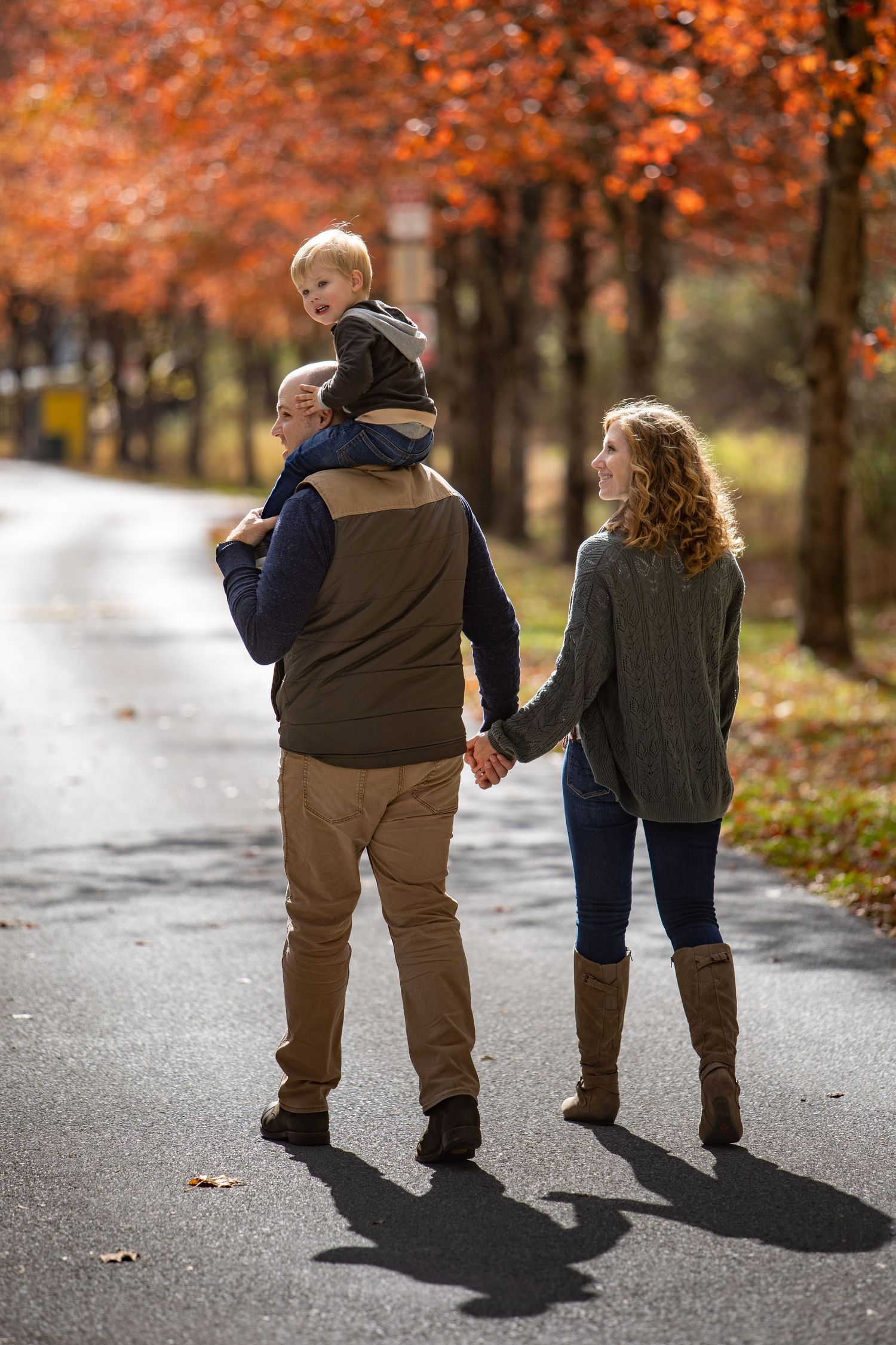 Two people walk hand in hand down a path surrounded by autumn trees with vibrant orange foliage casting long shadows.