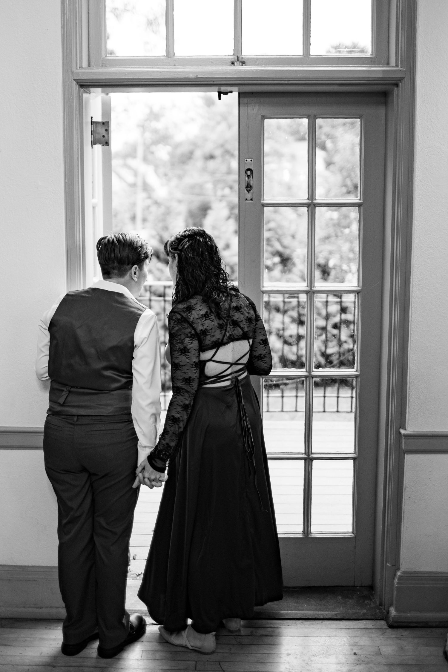 Black and white photo showing couple looking out window together in formal attire.