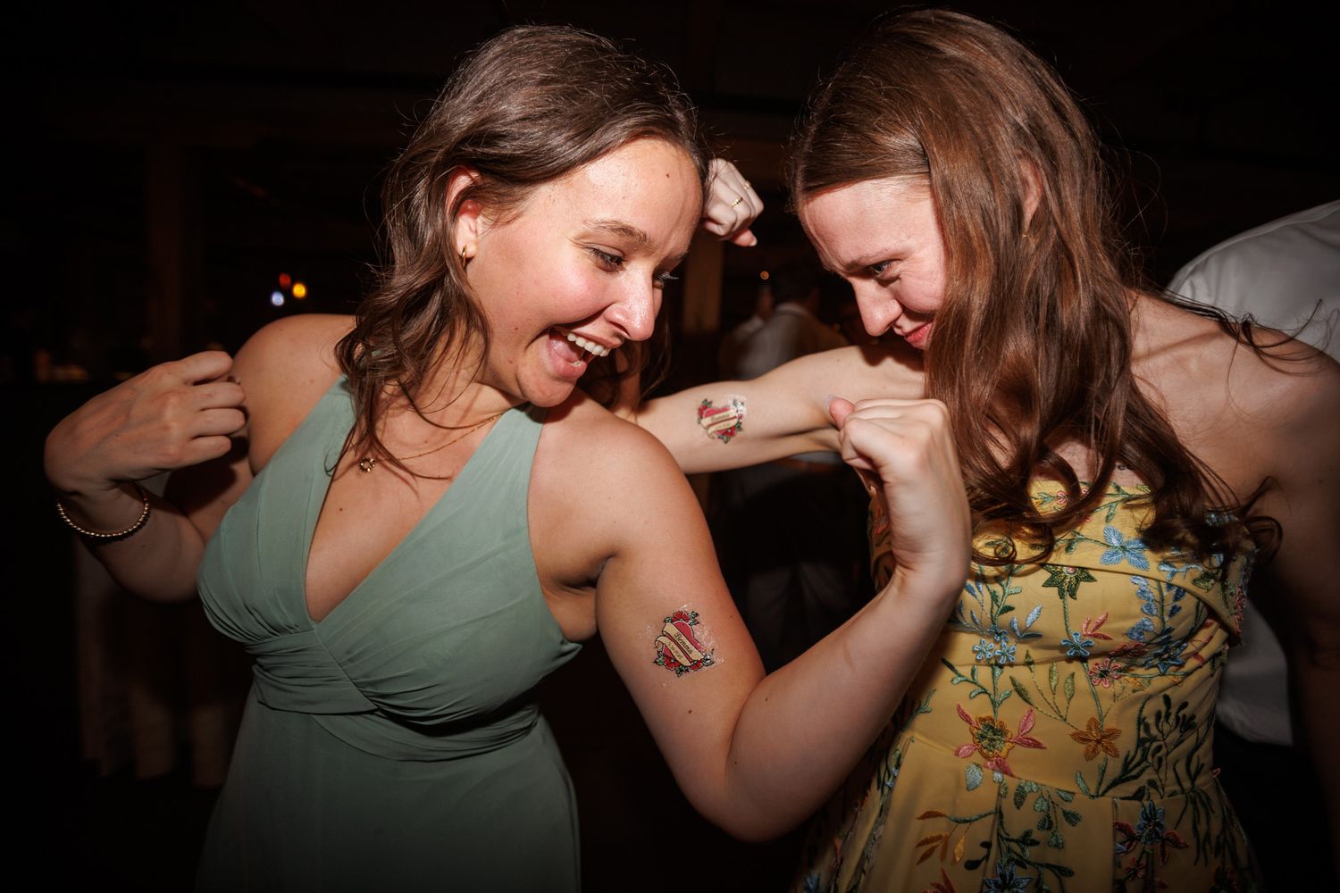 Two women laughing and comparing tattoos on their arms while dancing at a wedding reception.