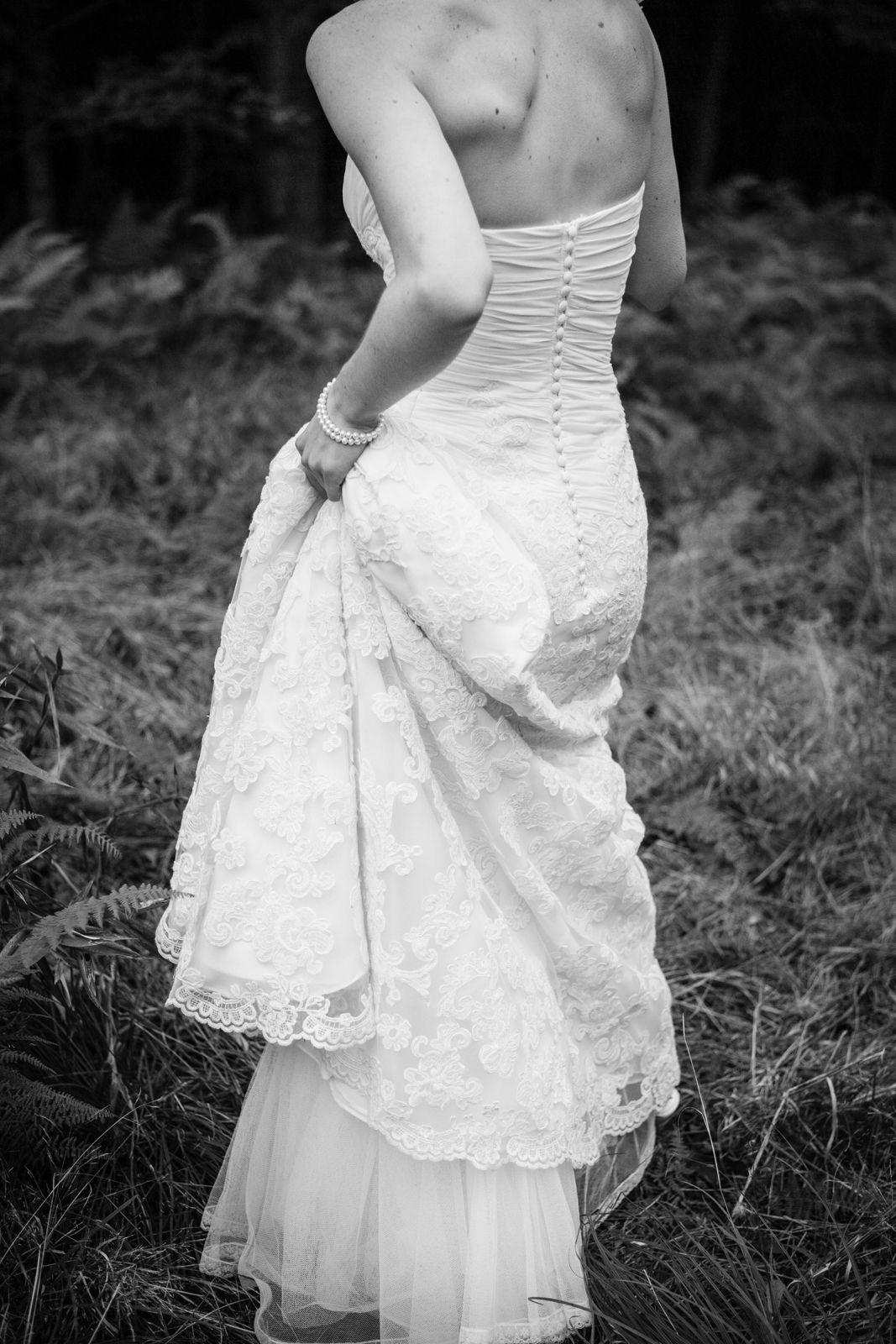 Black and white photo of a bride in a lace corset wedding gown holding her train in a natural outdoor setting.