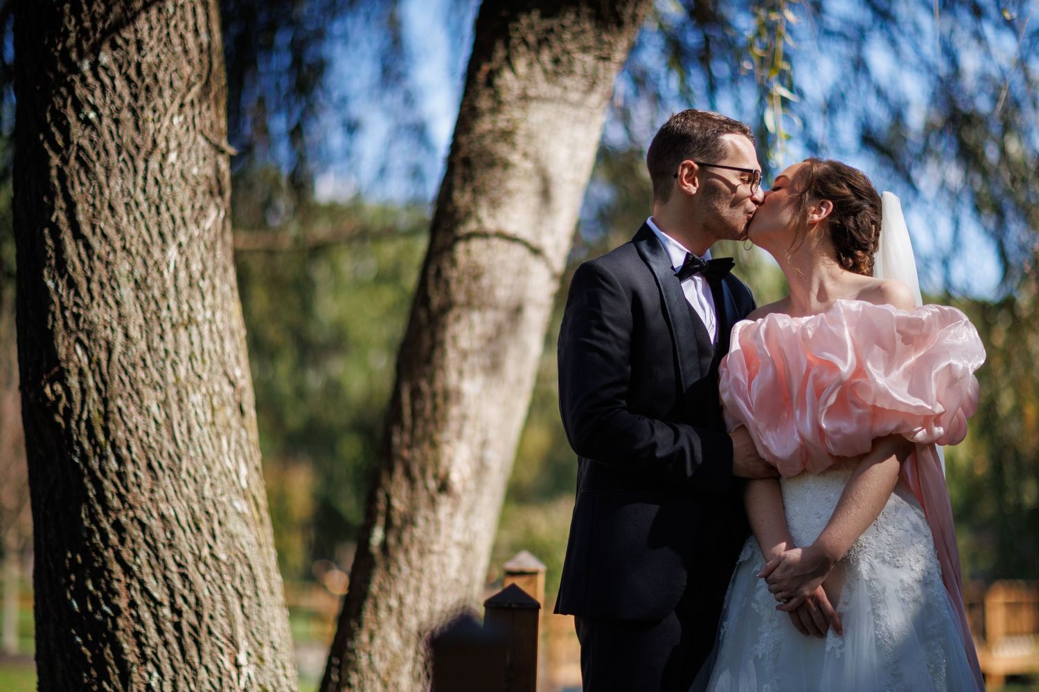 Bride and groom share a romantic kiss outdoors under a tree in wedding attire.