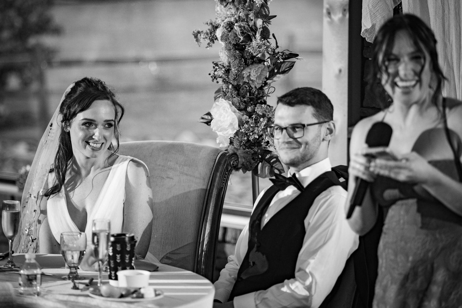 Bride and groom laugh joyfully during wedding speech at elegantly decorated reception table.