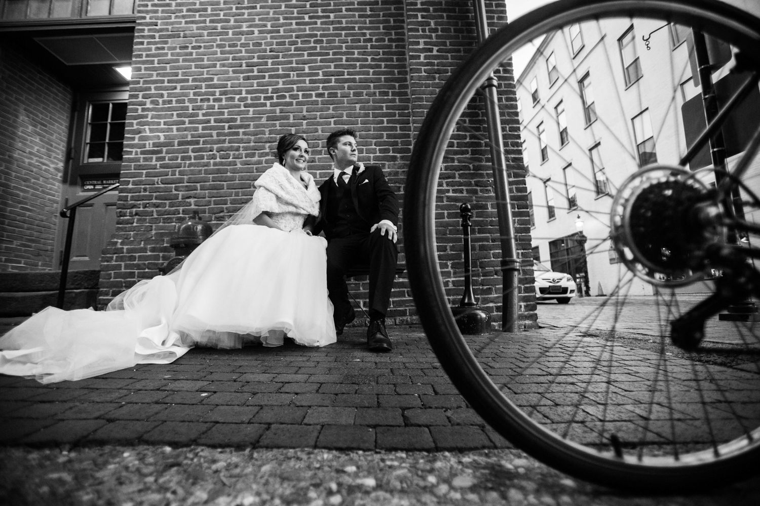 a wedding couple on a bench in Lancaster, PA central market. There is a bicycle wheel in the foreground. This is an unconventional portrait