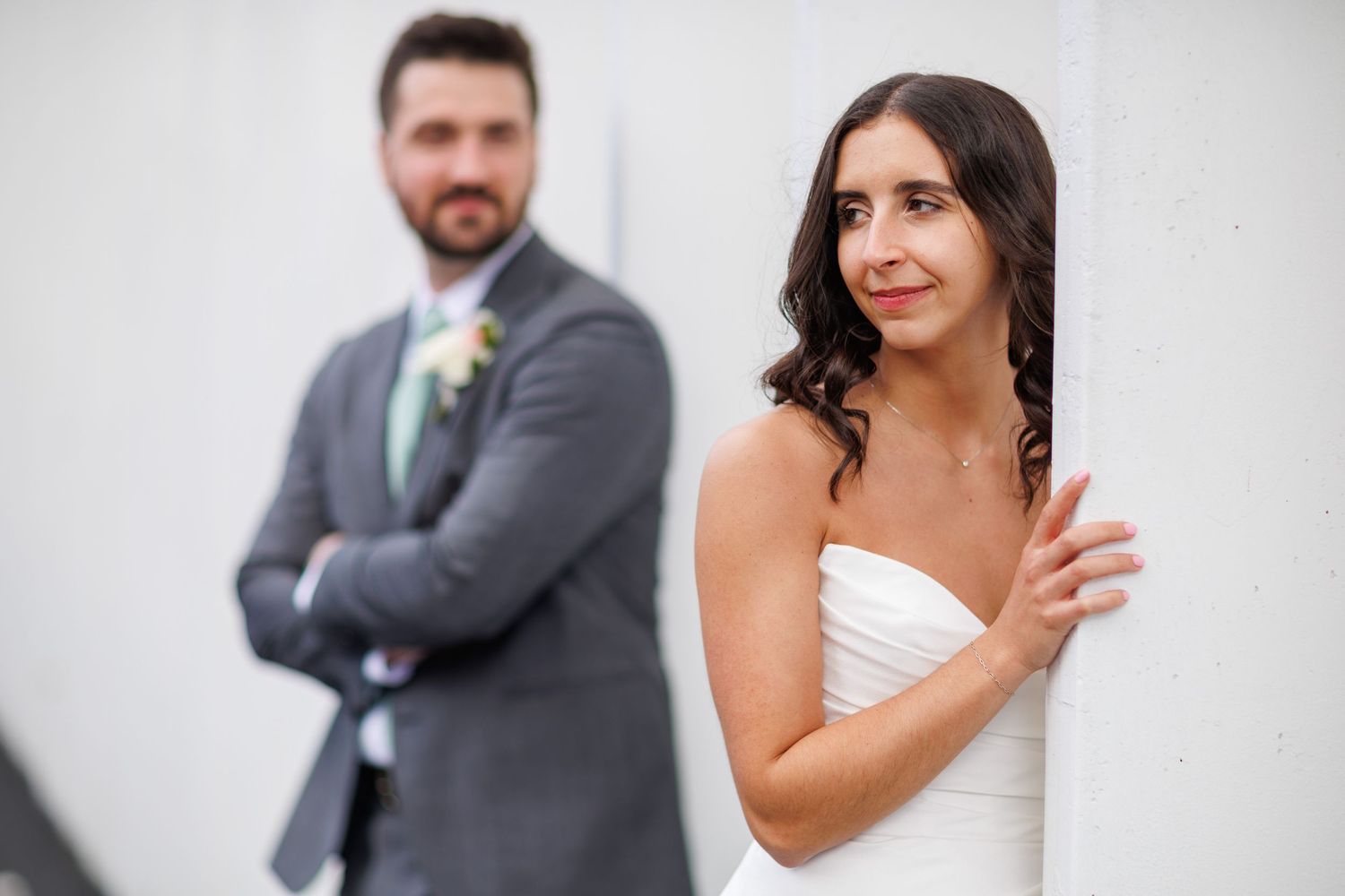 Bride in strapless white gown peeks around a white pillar while groom in gray suit stands behind her.