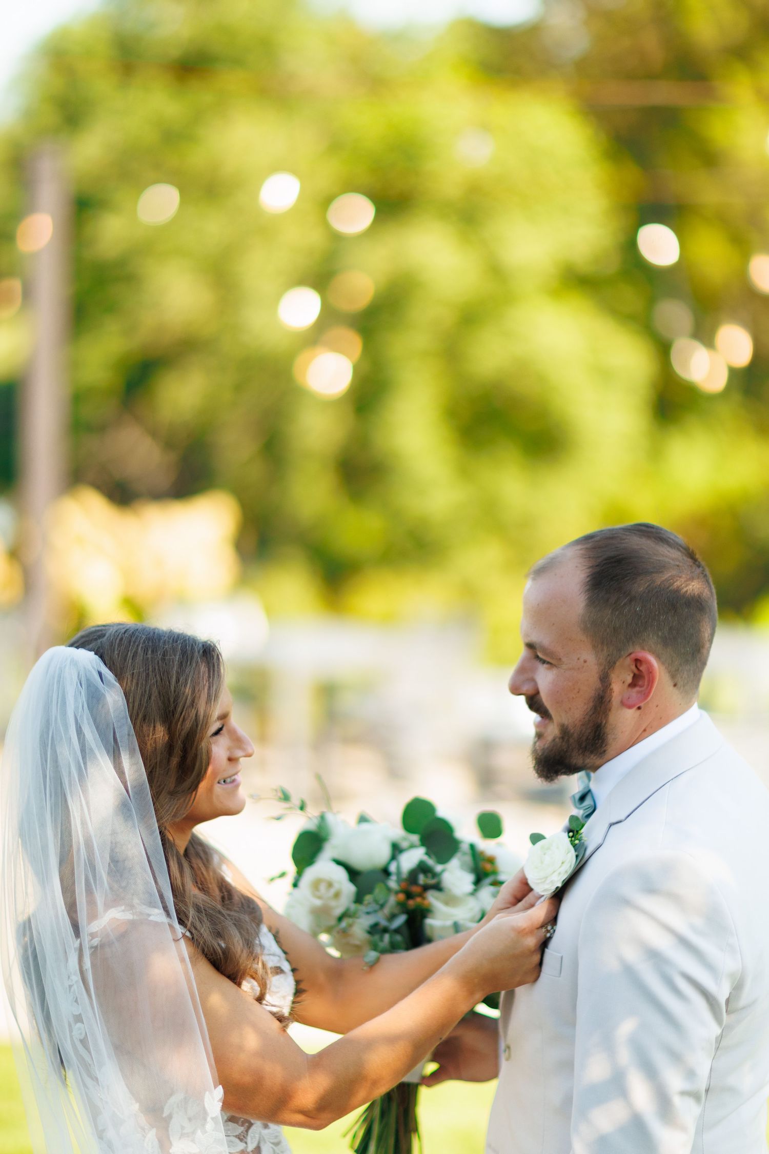 A newlywed couple shares a tender moment outdoors surrounded by soft bokeh lights and holding white and green flowers.