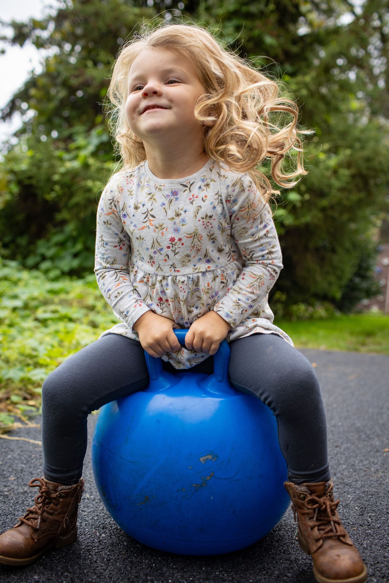 Child bouncing on blue exercise ball wearing brown boots and floral shirt.