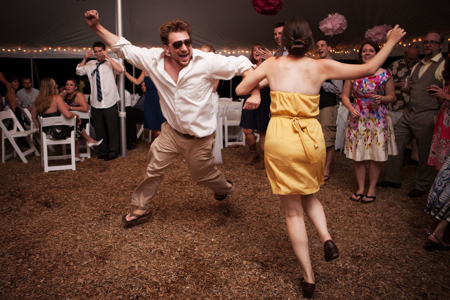 Energetic dancers in vintage attire perform enthusiastic moves at a rustic outdoor reception venue.