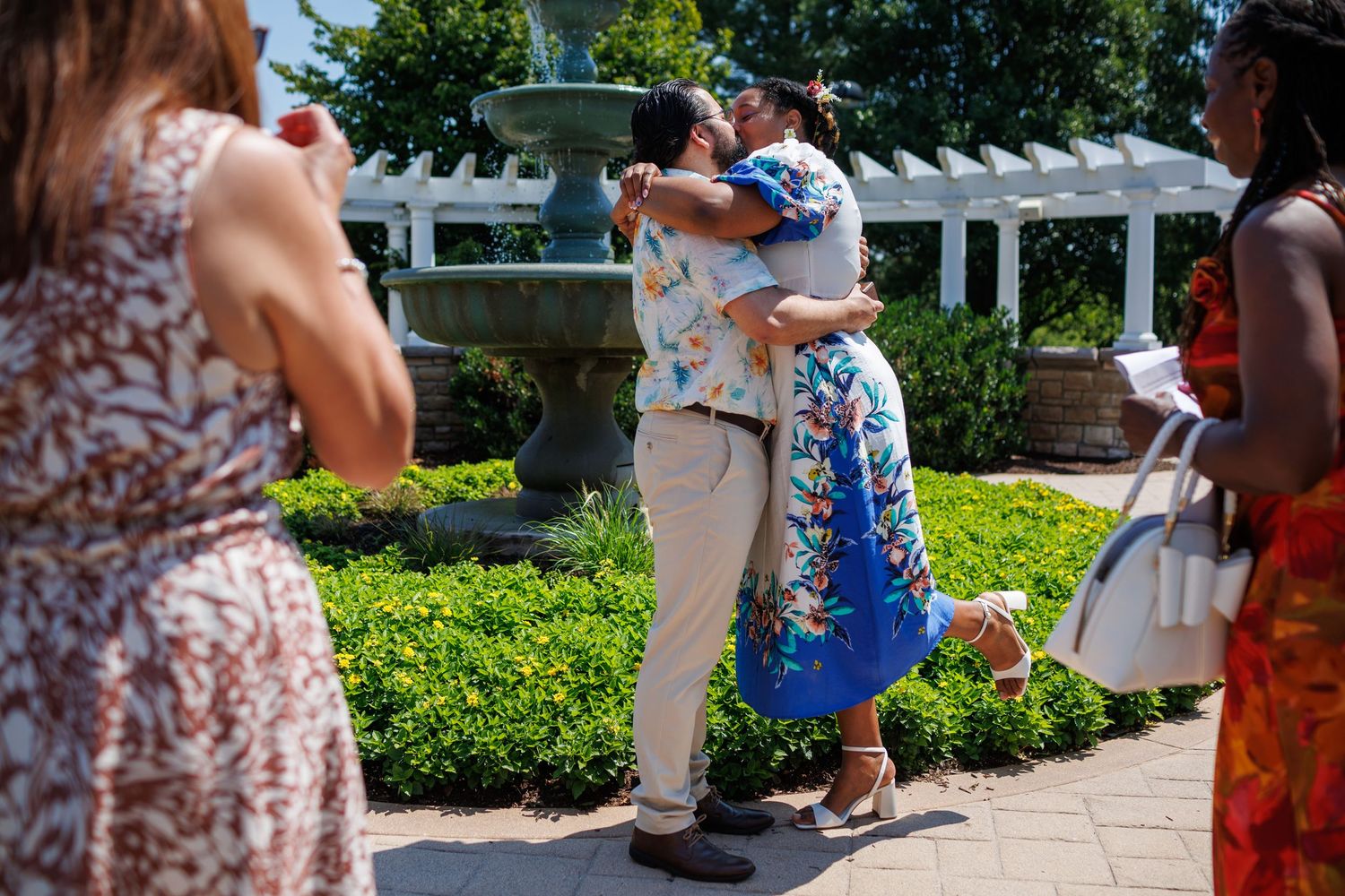 Couple embracing near a garden fountain at an outdoor event, with guests in floral attire surrounding them on a sunny day.