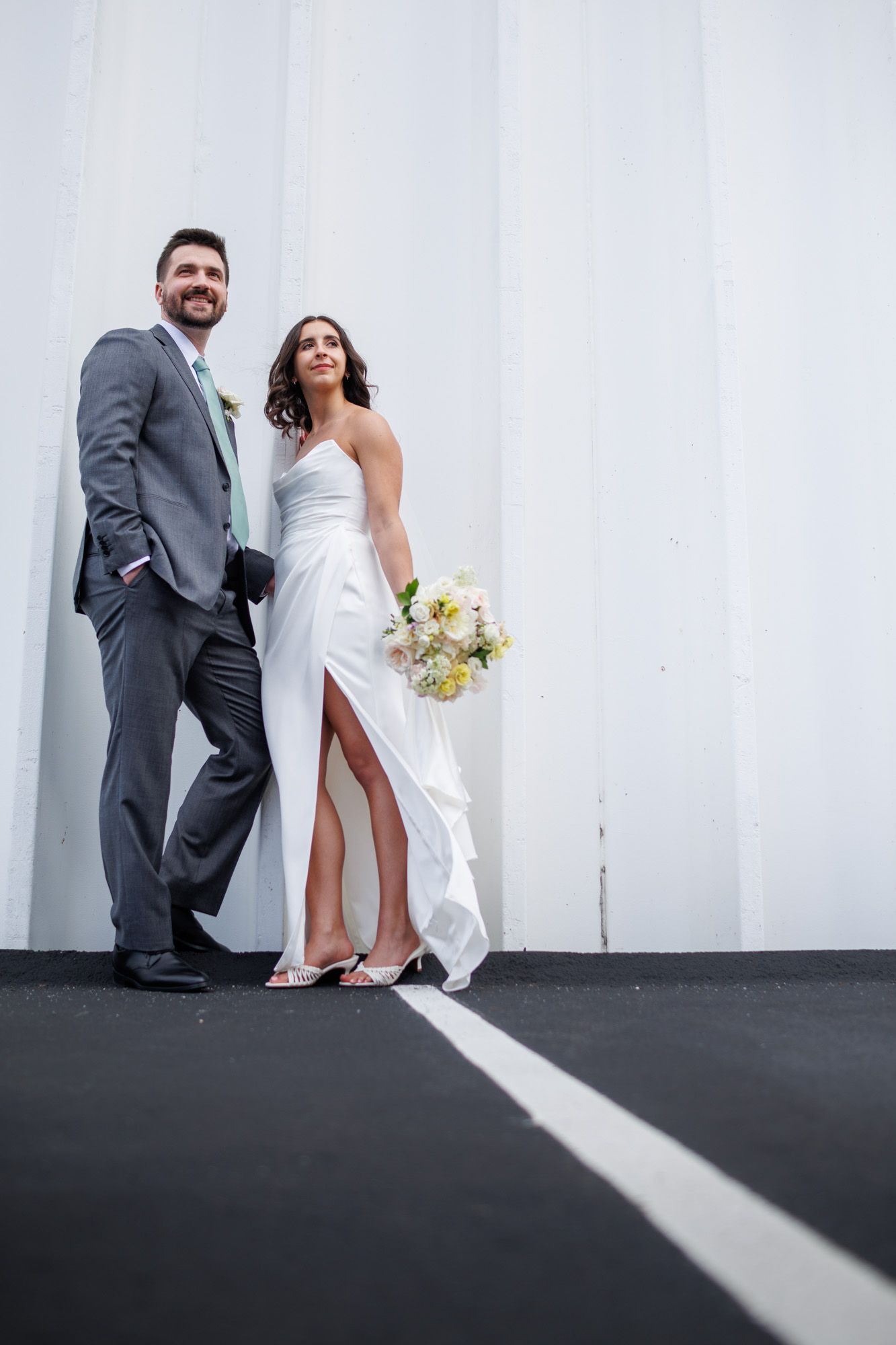 Smiling bride and groom pose against white wall, bride holding pastel bouquet in elegant white gown with slit.