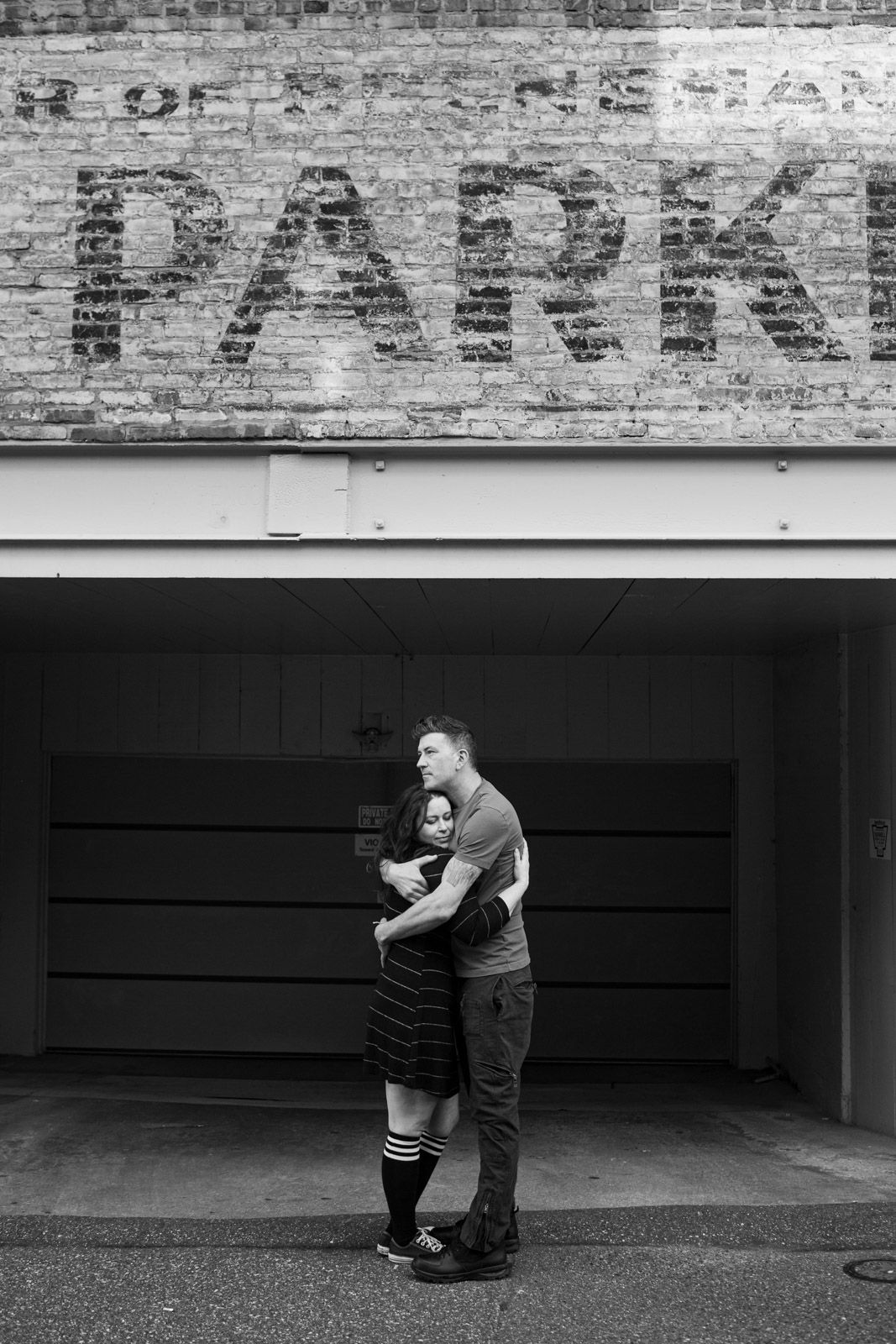 Black and white photo of a couple embracing in front of a brick wall with parking sign.
