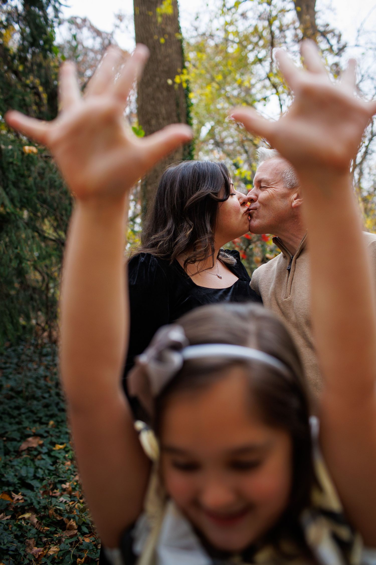 A playful outdoor family photo in autumn with raised hands in the foreground and a romantic moment in the background.