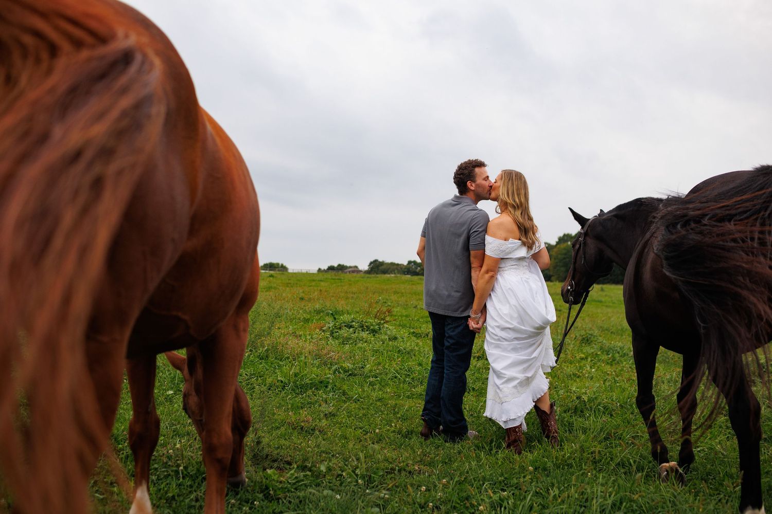 A couple shares a romantic kiss in a green field surrounded by horses on a cloudy day.
