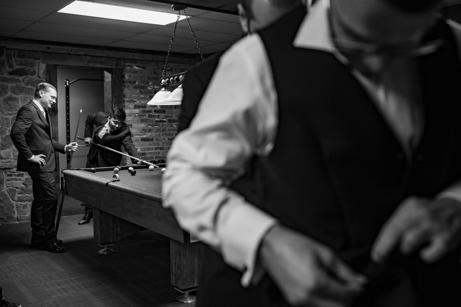 Black and white photo of men in formal attire playing pool in a rustic billiard room during a wedding.