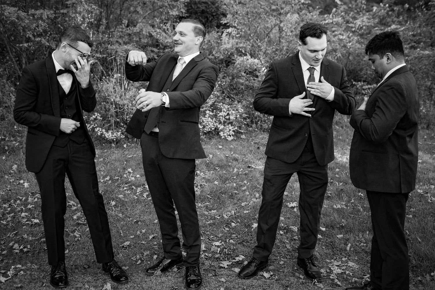 Four groomsmen in black suits share drinks and laugh outdoors at a wedding celebration.