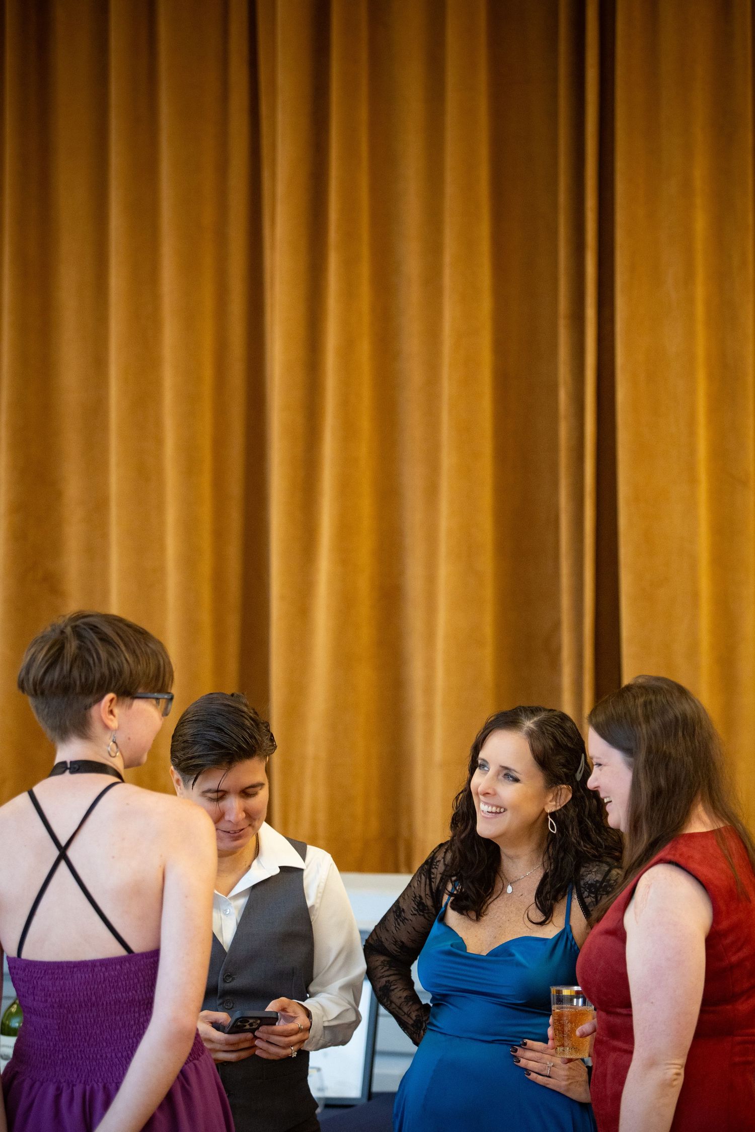 Group of people in formal attire socializing against golden curtain backdrop.