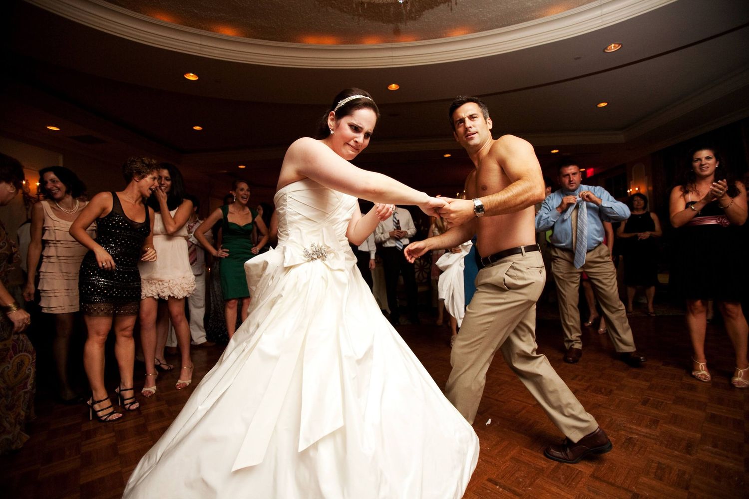 A dancing couple spins energetically on a wooden dance floor while wedding guests watch from the sides during a reception.