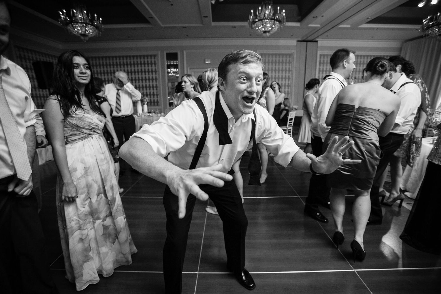 Party guests dance energetically at a wedding reception under crystal chandeliers in black and white photography.