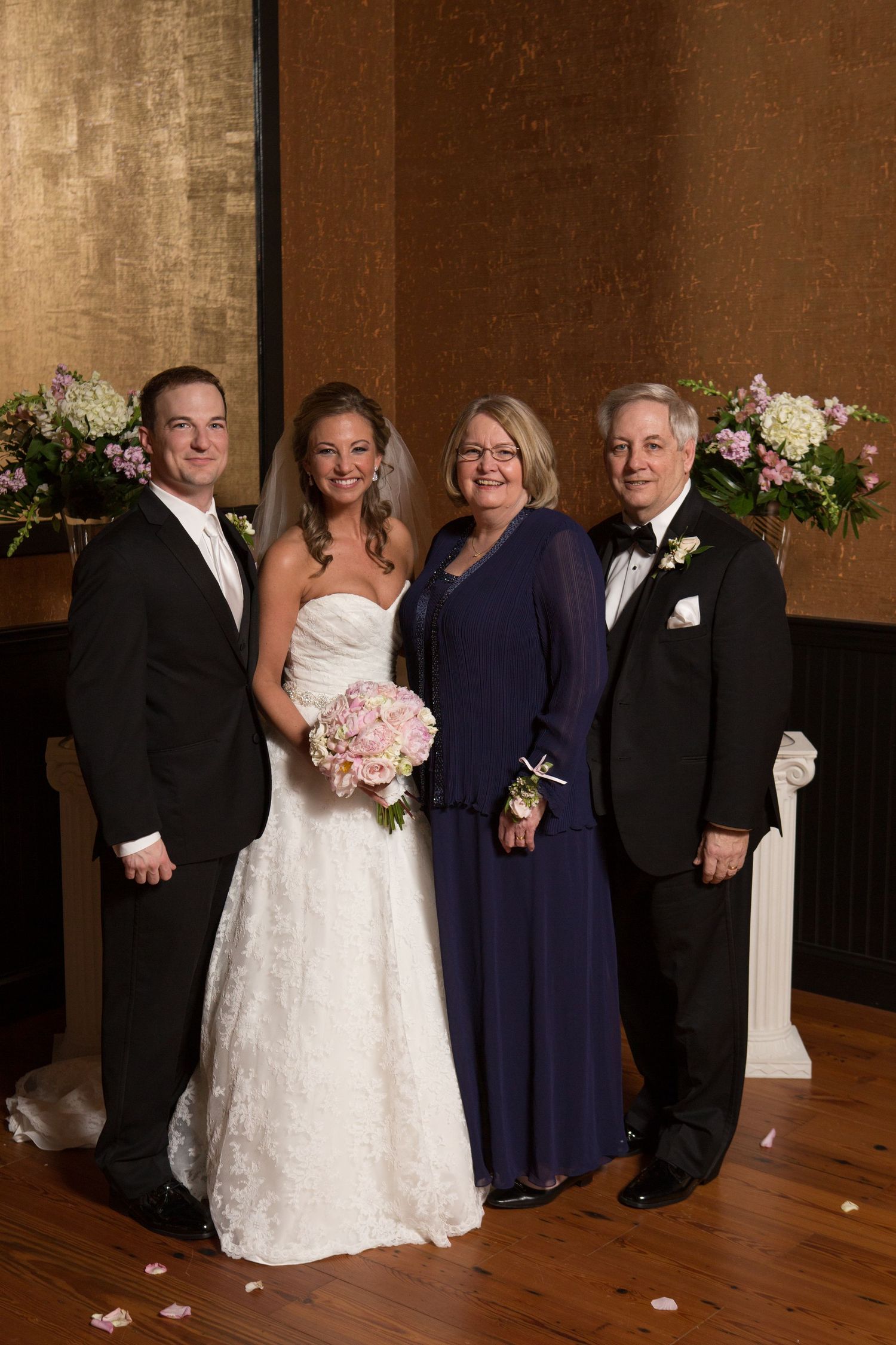 Group photo of wedding party with bride in white dress and others in formal navy and black attire against brown backdrop.