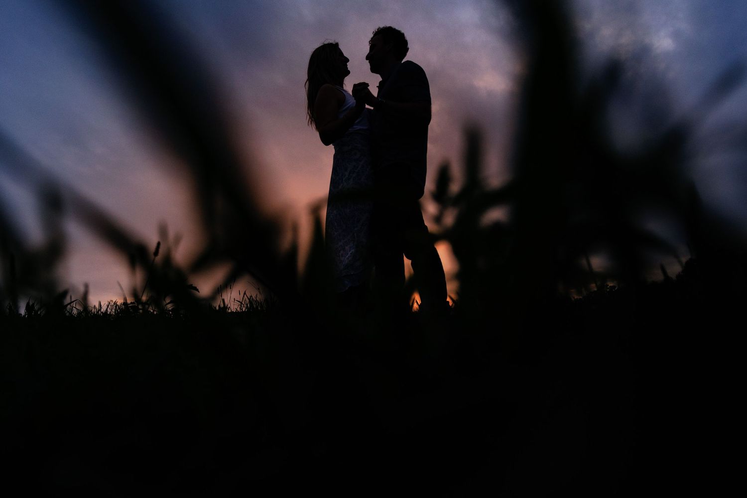 Silhouette of a romantic couple embracing at twilight with a colorful sunset sky and dark foreground grass.