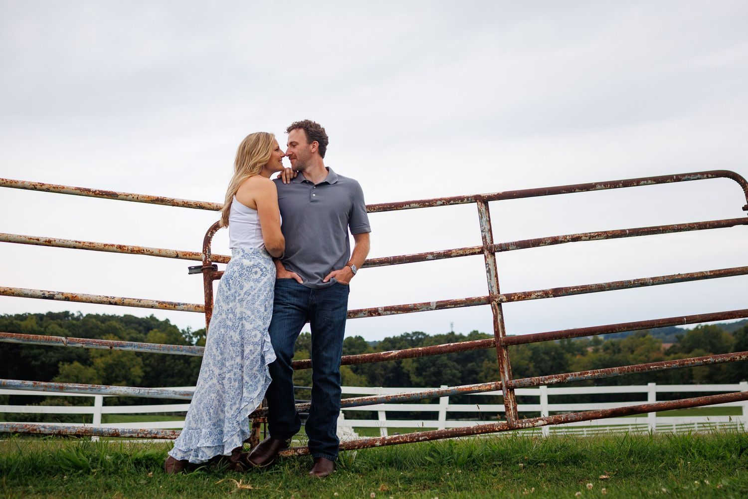 Couple standing together by rustic metal farm gate on green countryside, sharing a romantic moment outdoors.