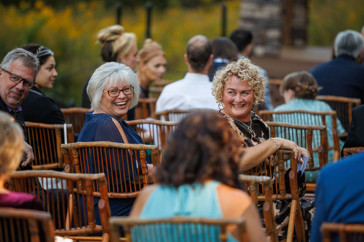 Guests laughing and socializing at an outdoor wedding ceremony seated in wooden chairs.