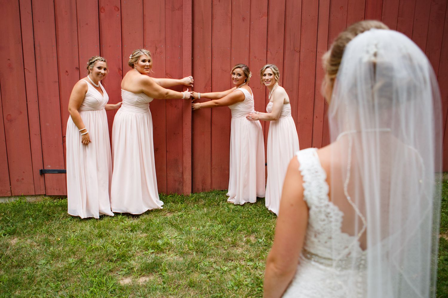 A bridal party in soft pink dresses shares a playful moment in front of a red barn wall while the bride looks on.