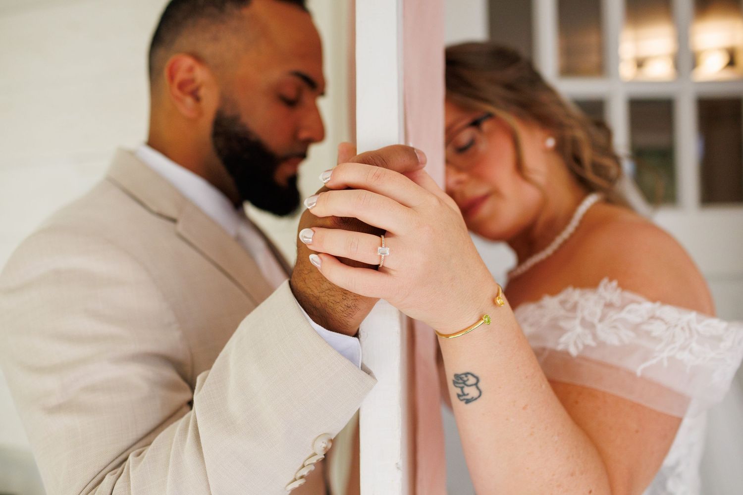 Bride and groom lean against a door, hands intertwined, showcasing wedding rings and a delicate gold bracelet.