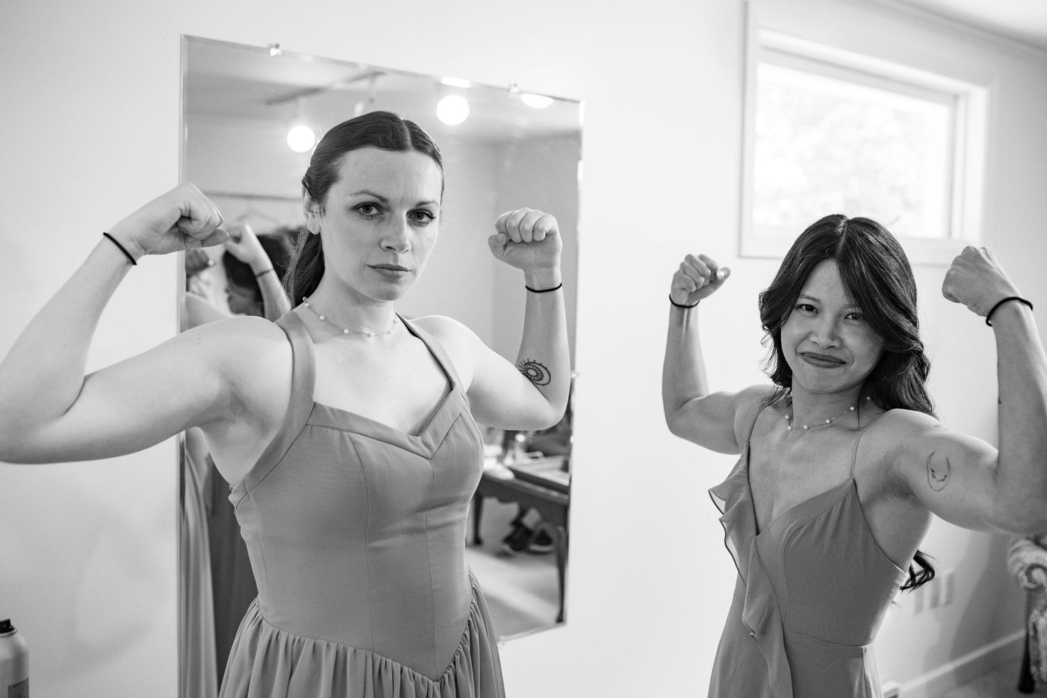 Two bridesmaids in gray dresses flex their muscles playfully in front of a mirror in black and white photo.