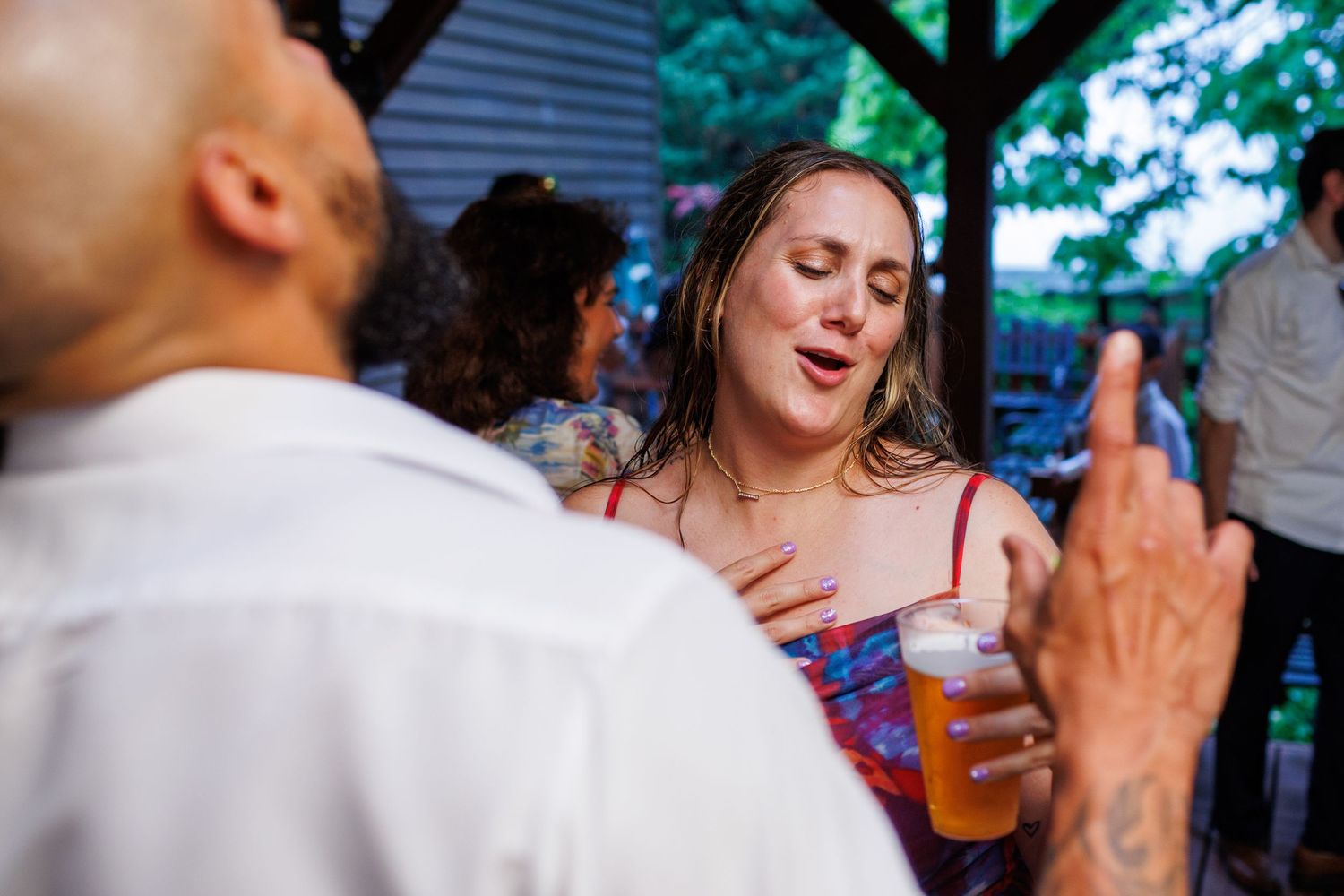 Wedding guest in red dress holds beer and chats at lively outdoor reception party.