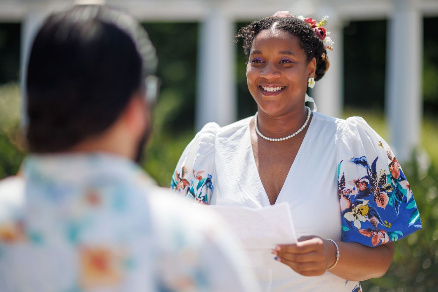 A smiling Black woman in a white floral dress holds papers outdoors near white columns on a sunny day.