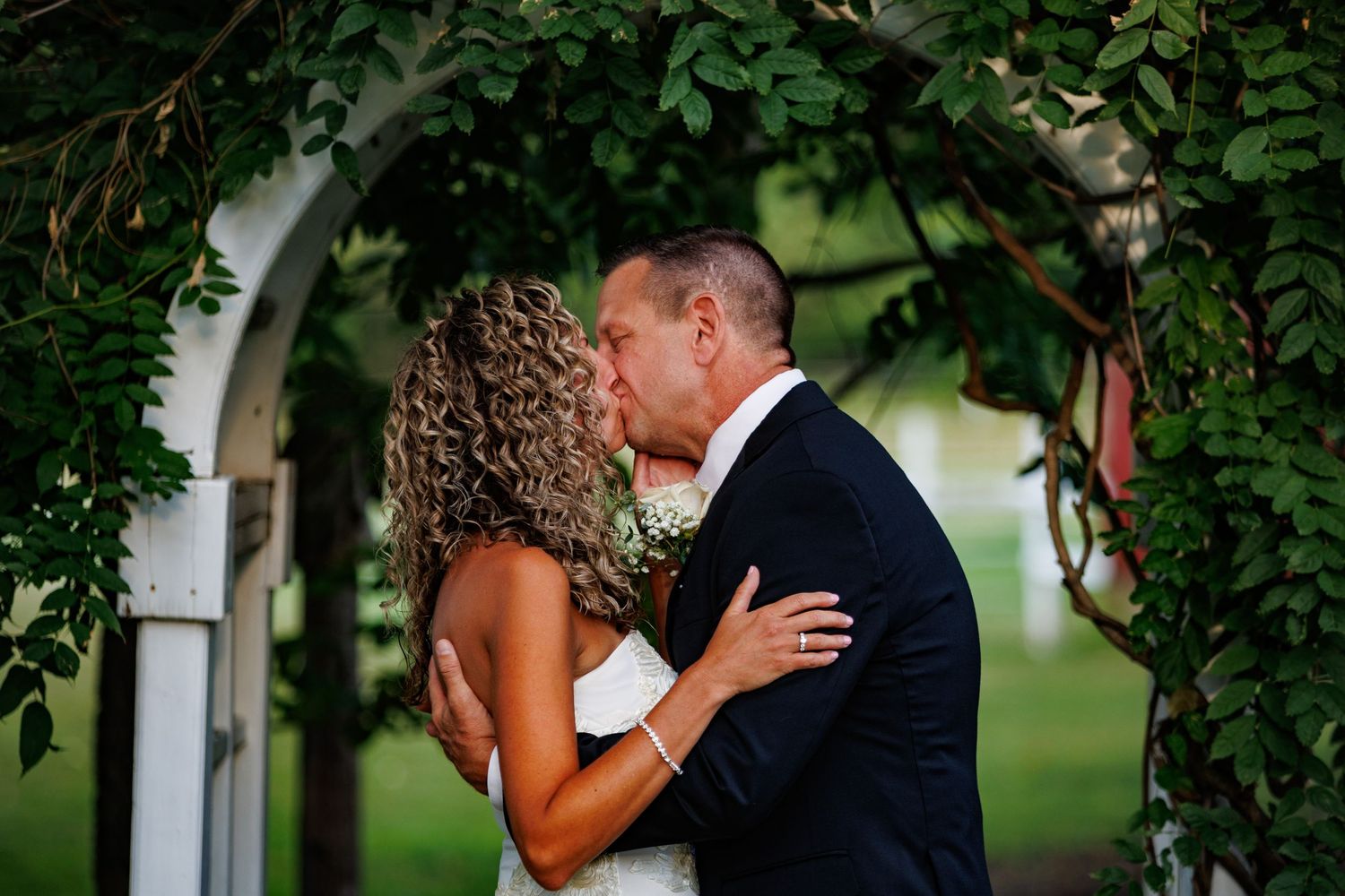 Newlyweds share a romantic kiss under a lush green floral arch, bride in white gown and groom in navy suit.