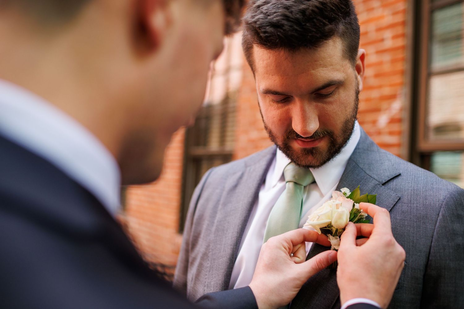 Groomsman pinning a white boutonniere on groom in gray suit and green tie before wedding ceremony.