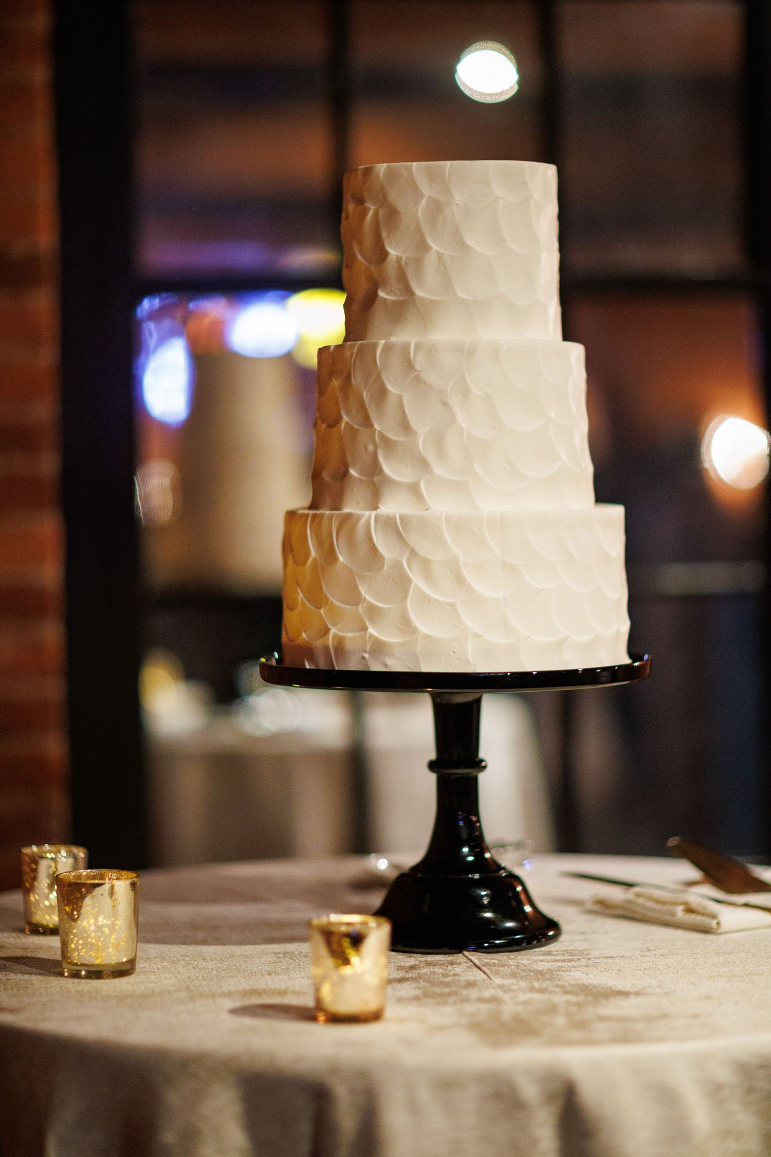 Elegant four-tier white wedding cake with petal texture on a black stand surrounded by gold candles.