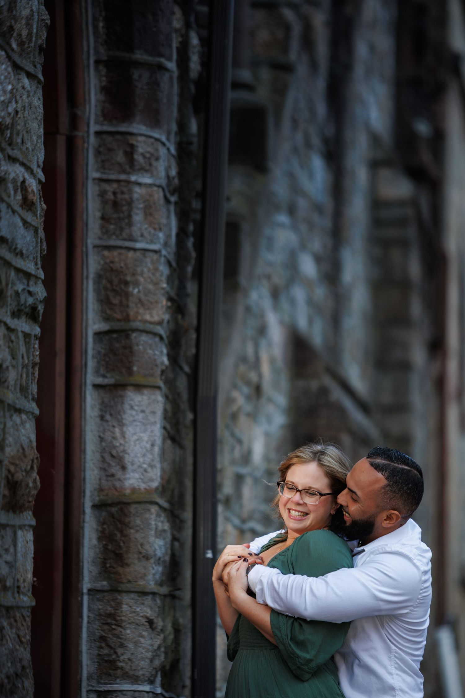 A romantic couple embraces against a stone wall with arched windows creating an intimate architectural backdrop.