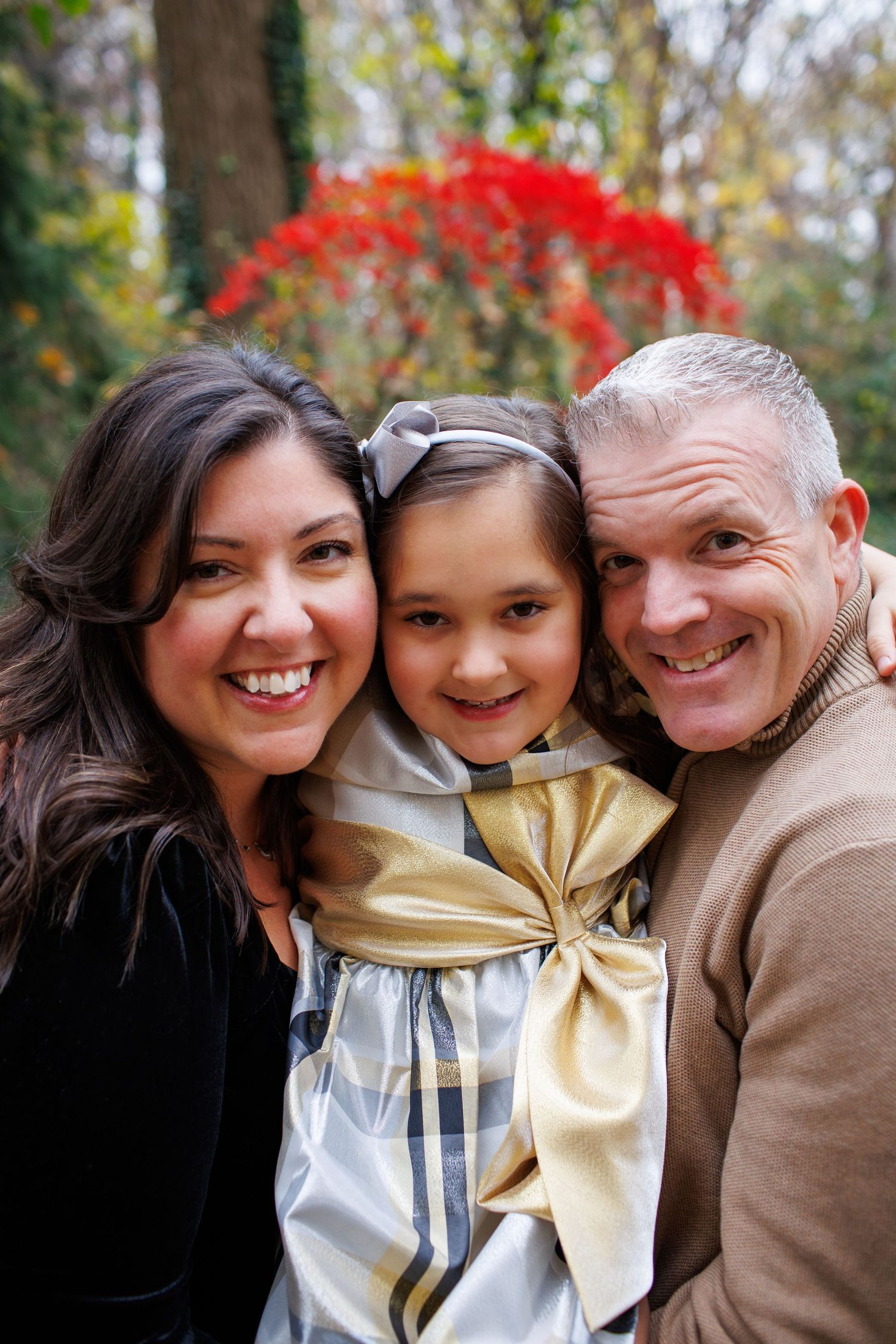 A family poses together outdoors with autumn foliage and red maple leaves visible in the background.