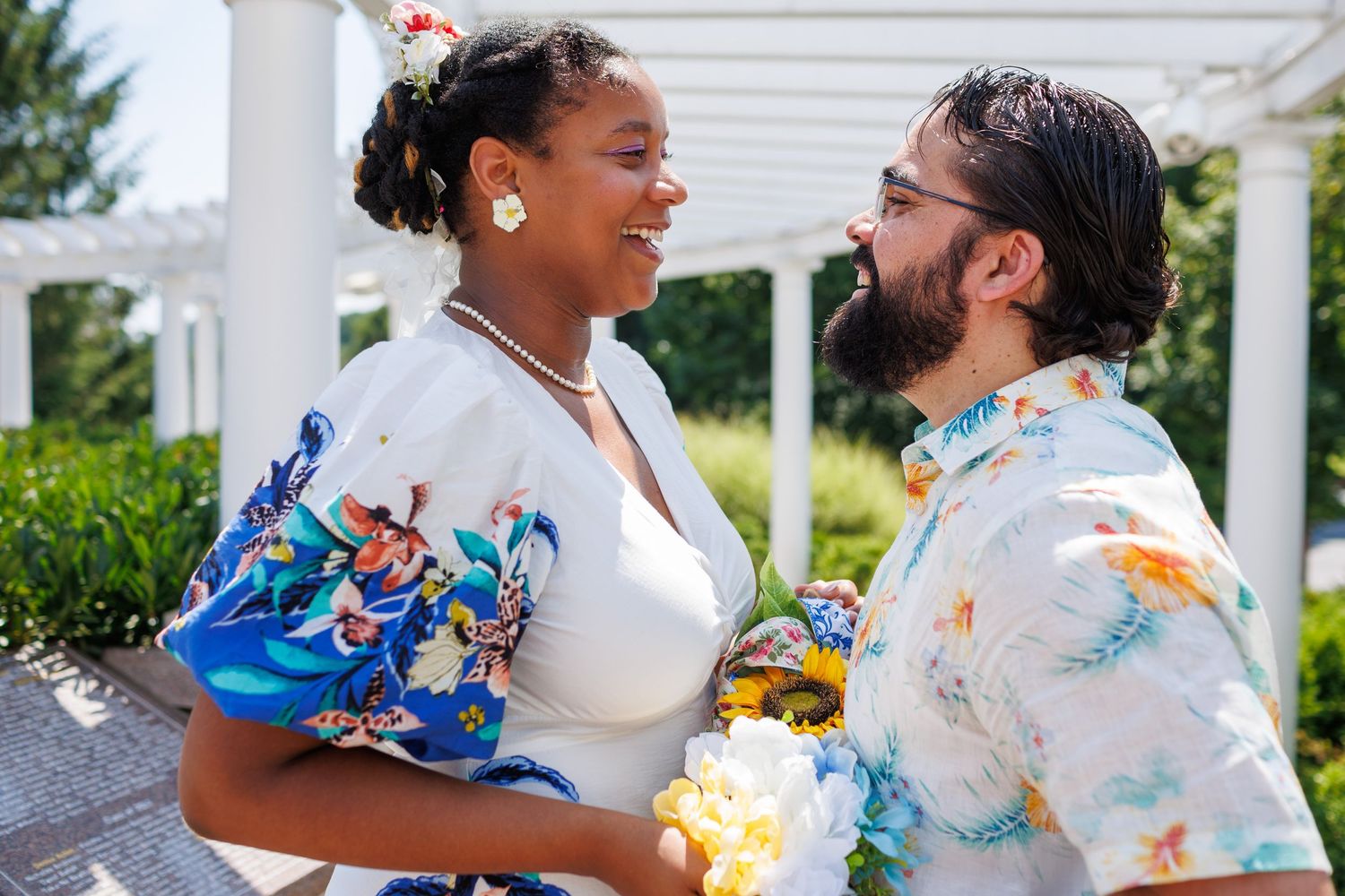 Happy couple in coordinating floral outfits smiling at each other near white pergola outdoors.
