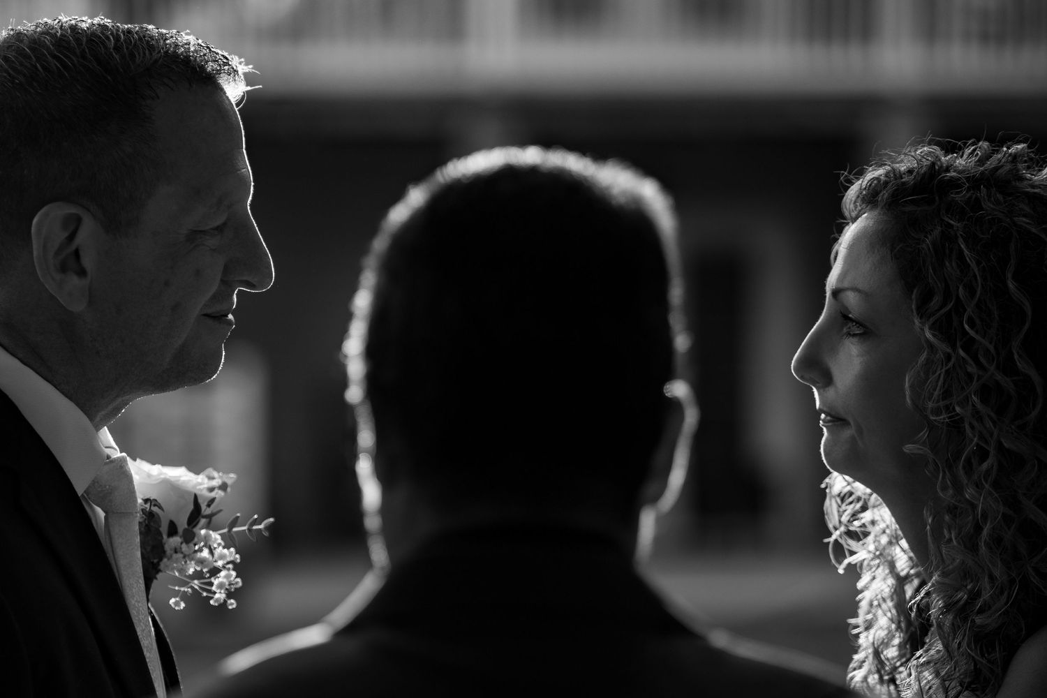 Black and white wedding ceremony photo of bride and groom facing officiant, captured in dramatic contrast lighting.