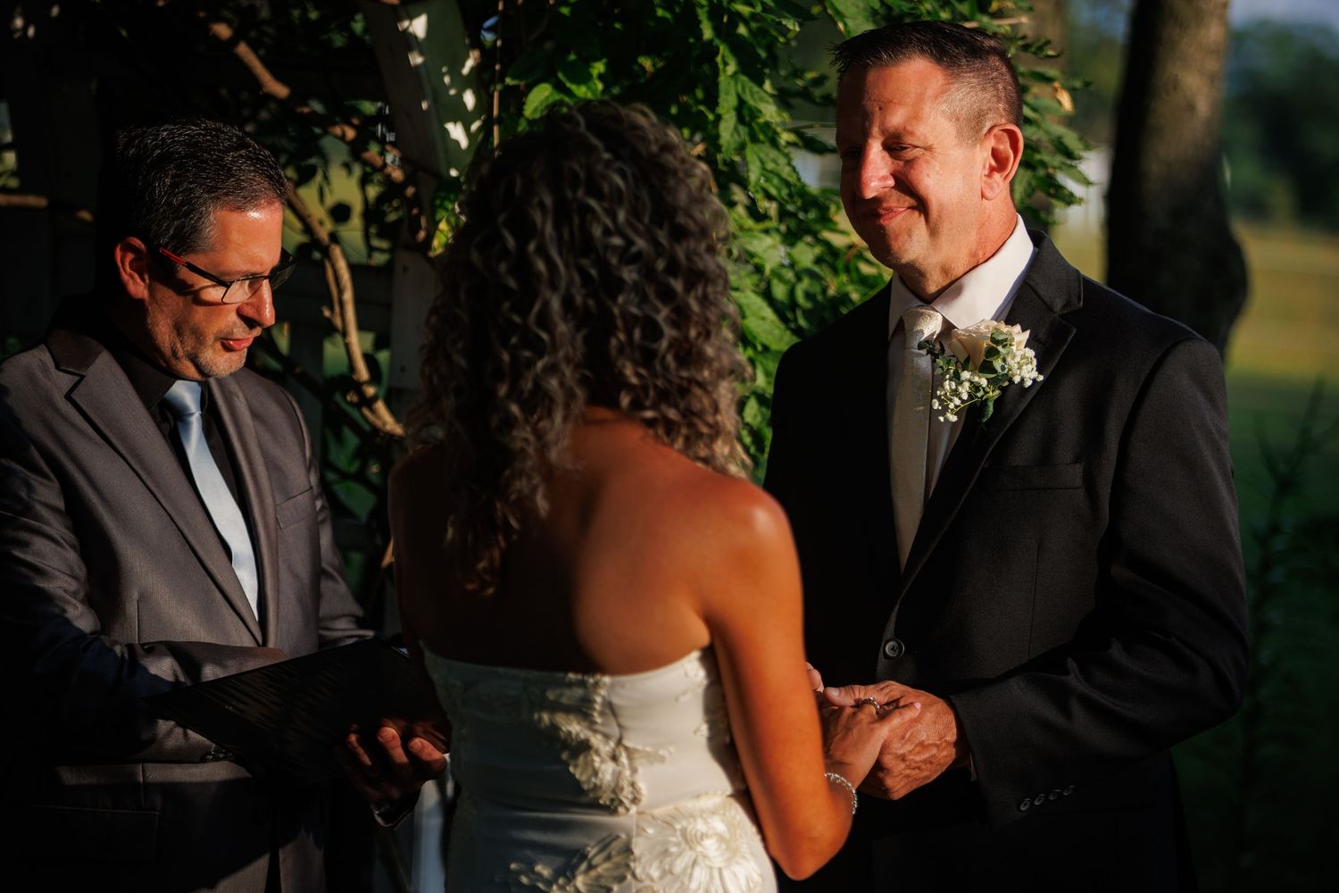 Groom smiling joyfully during outdoor wedding ceremony while exchanging rings with bride before officiant.