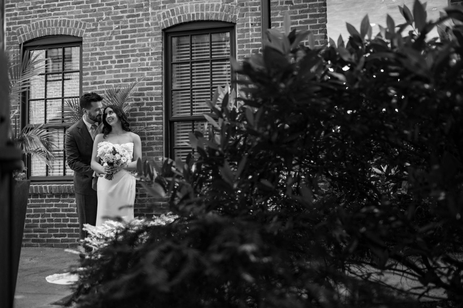 Black and white photo of bride and groom embracing beside brick building surrounded by lush greenery.
