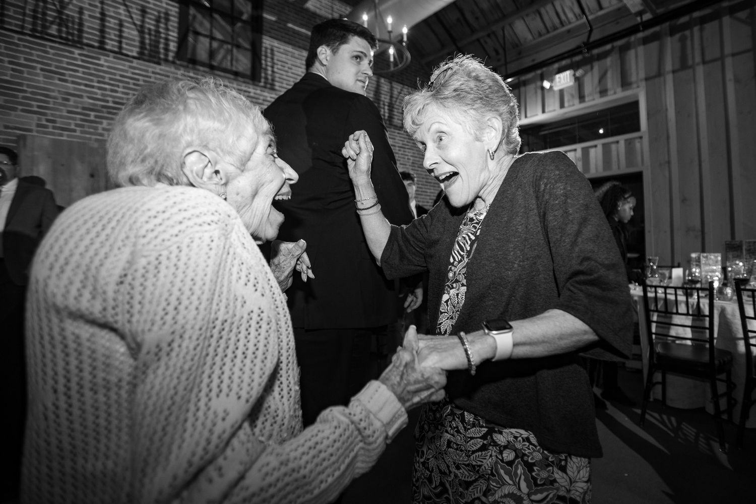 Two elderly women laughing and holding hands while dancing at an indoor wedding reception.