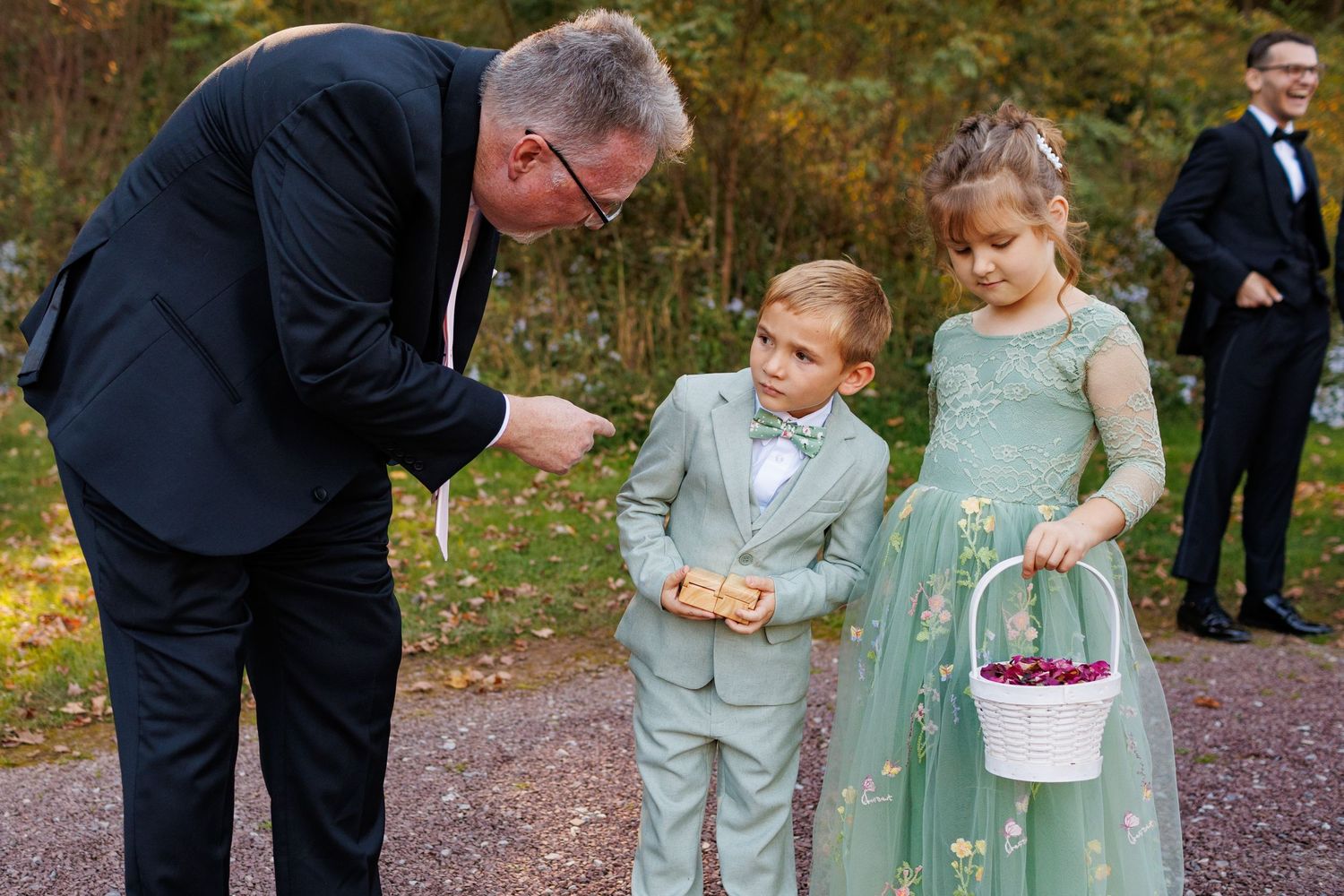 Man in formal suit interacts with young ring bearer and flower girl in sage green dress at outdoor wedding.