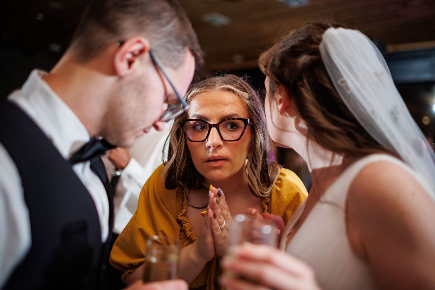 A woman with glasses leans in curiously between the bride and groom at a wedding.