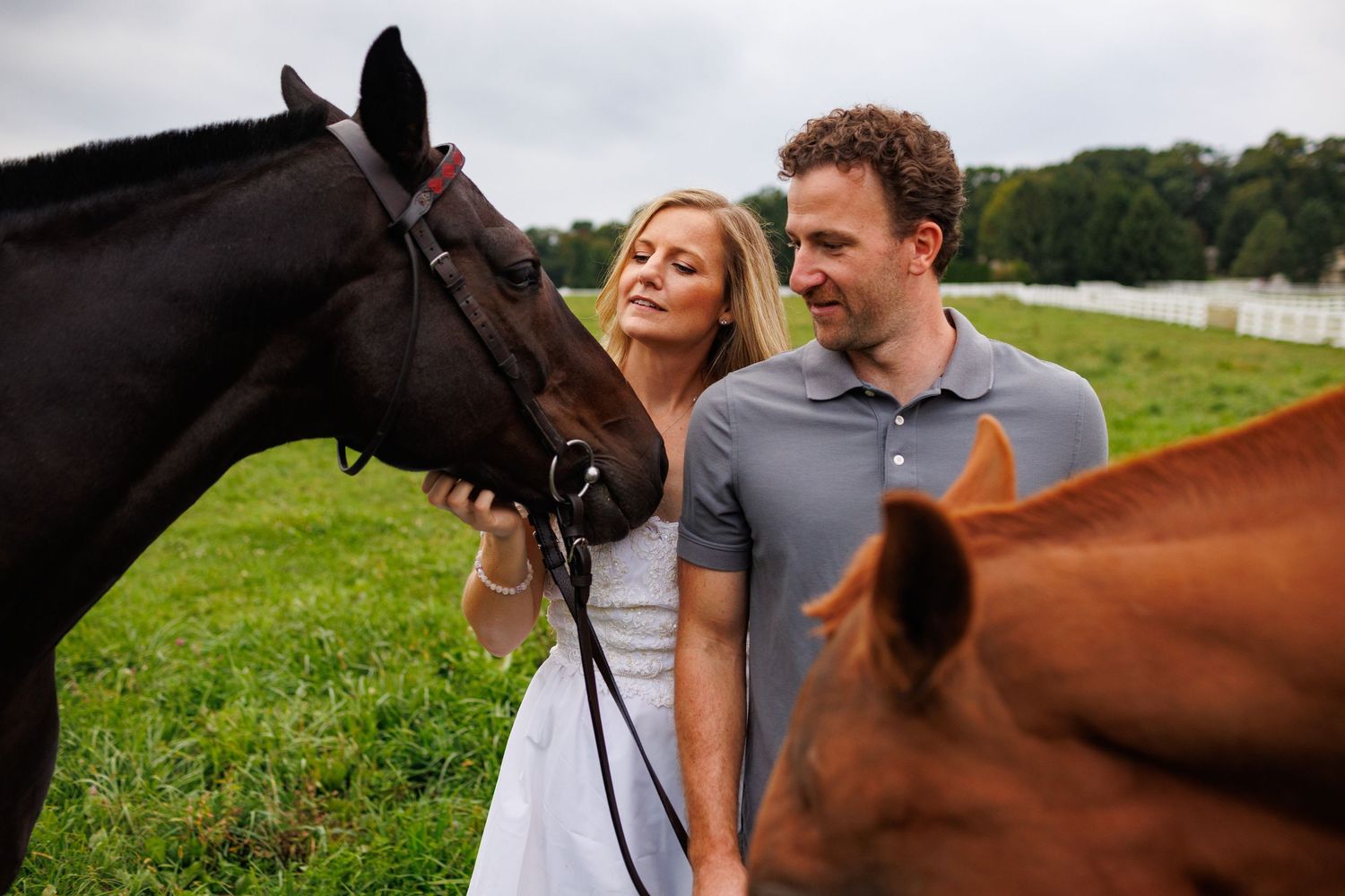 Couple smiling while petting a dark horse in a green field with a brown horse nearby.